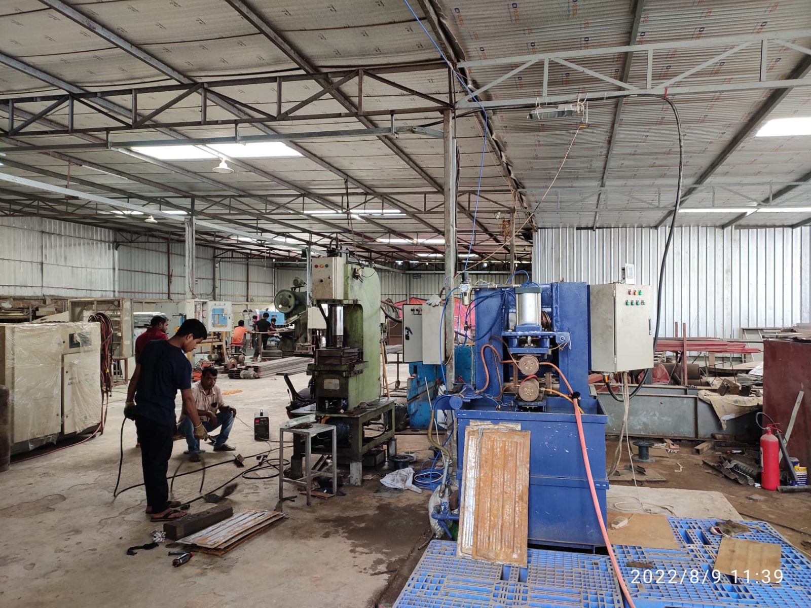Large factory workshop with hydraulic presses, a blue forming machine and workers handling sheet-metal parts