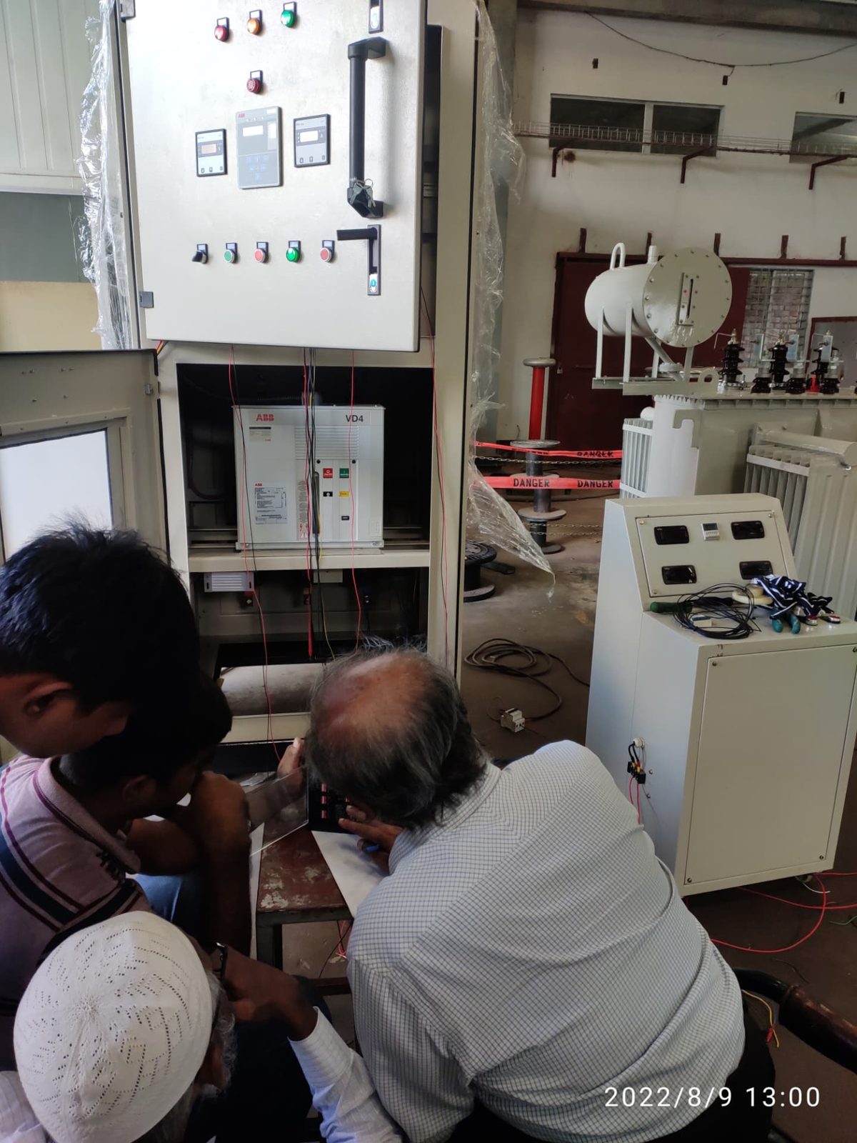 Engineers working at an open medium-voltage switchgear panel with an ABB VD4 vacuum circuit breaker, a test set and a distribution transformer behind