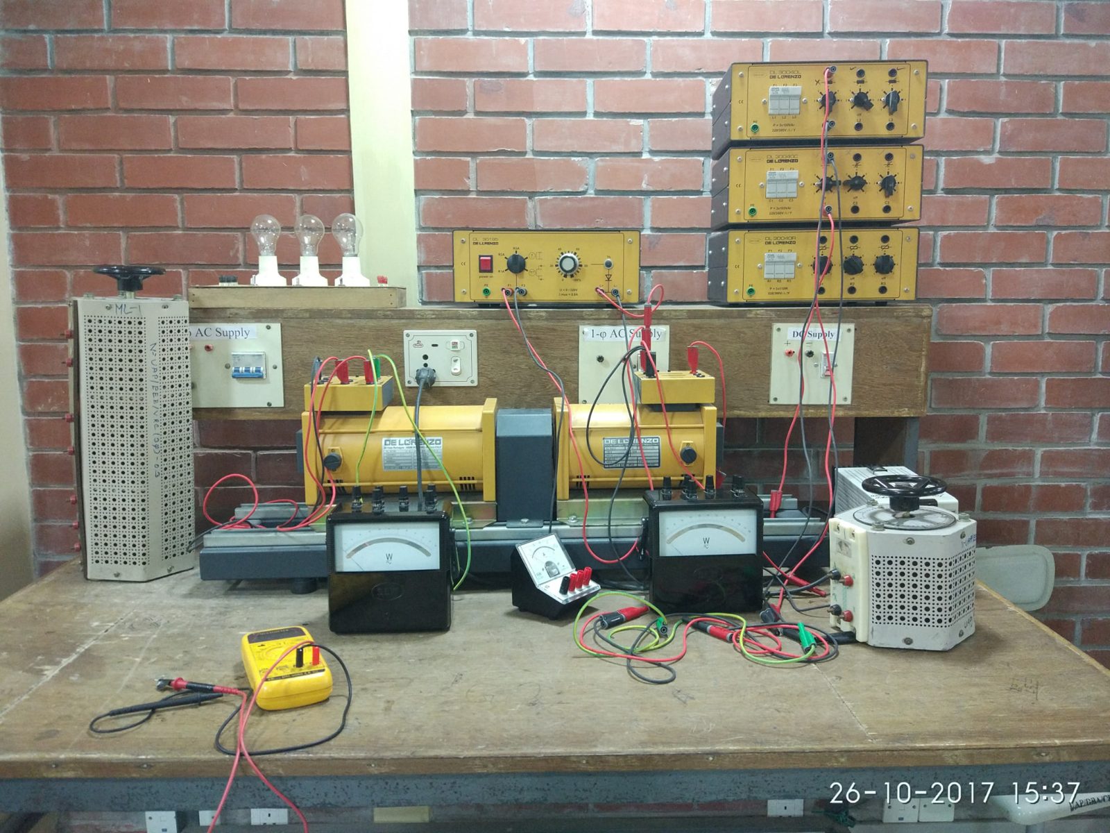 Lab bench with a coupled motor-generator set, wattmeters, an ammeter, variacs, load modules and a digital multimeter