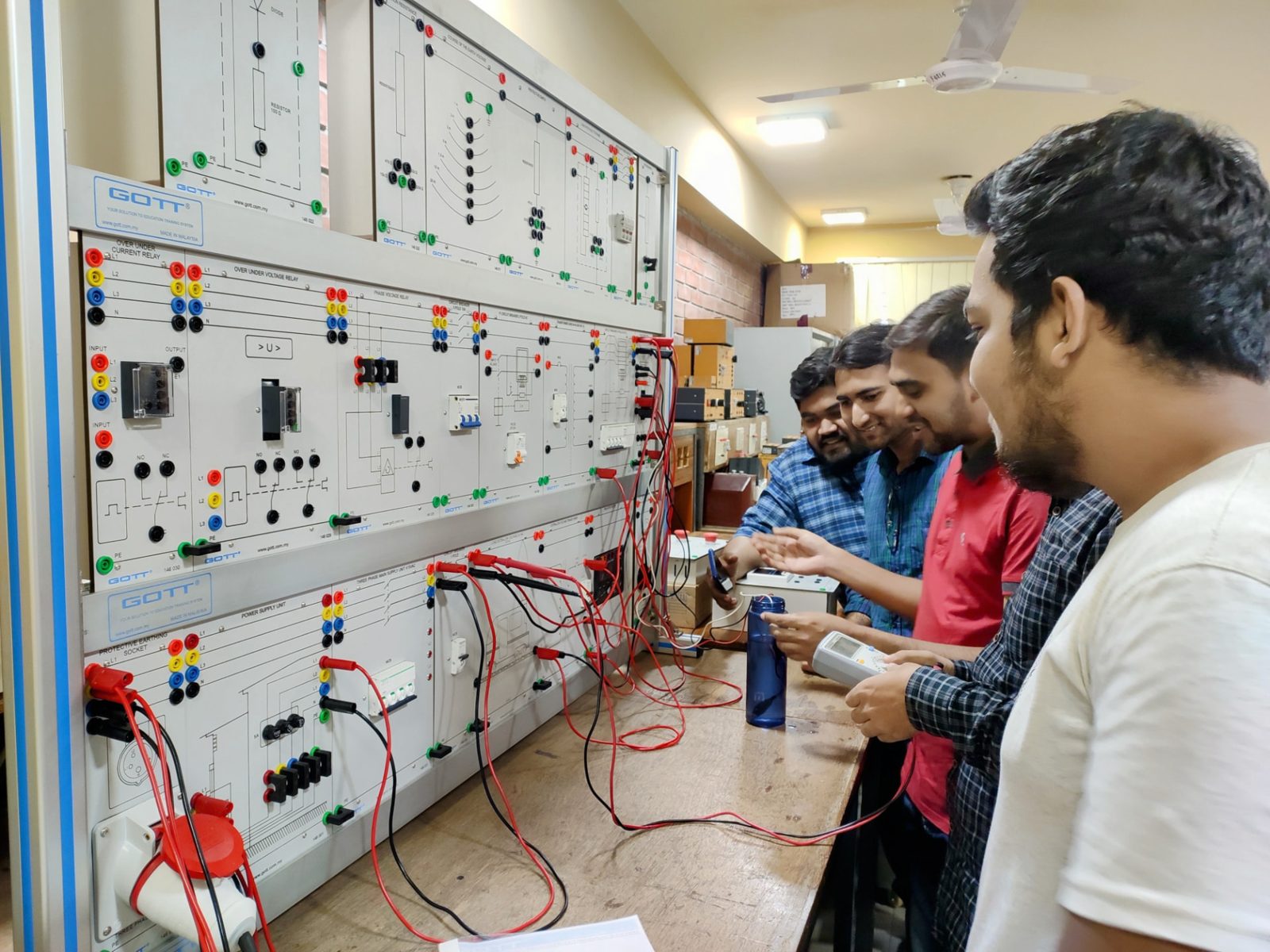 Group of engineers testing a protection relay training panel with over/under current and voltage relays, using a handheld meter