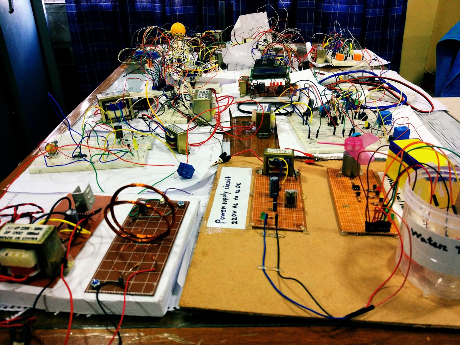 Table full of student electronics projects: a 220 V AC to 12 V DC power supply board, coils, transformers, relays and breadboard circuits